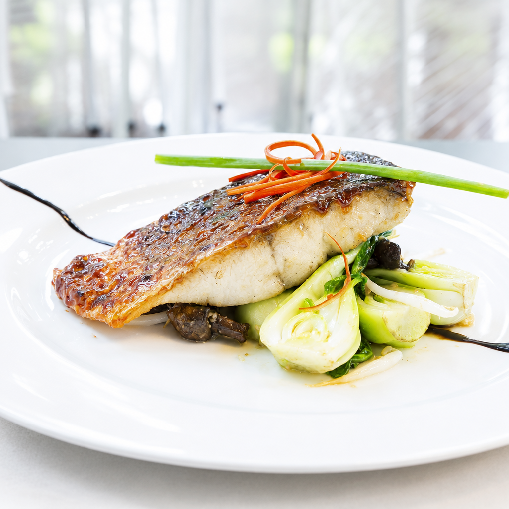 A plated cooked fish fillet with a seared top, served over bok choy and mushrooms on a white plate, with green and orange garnish in a softly blurred indoor setting.